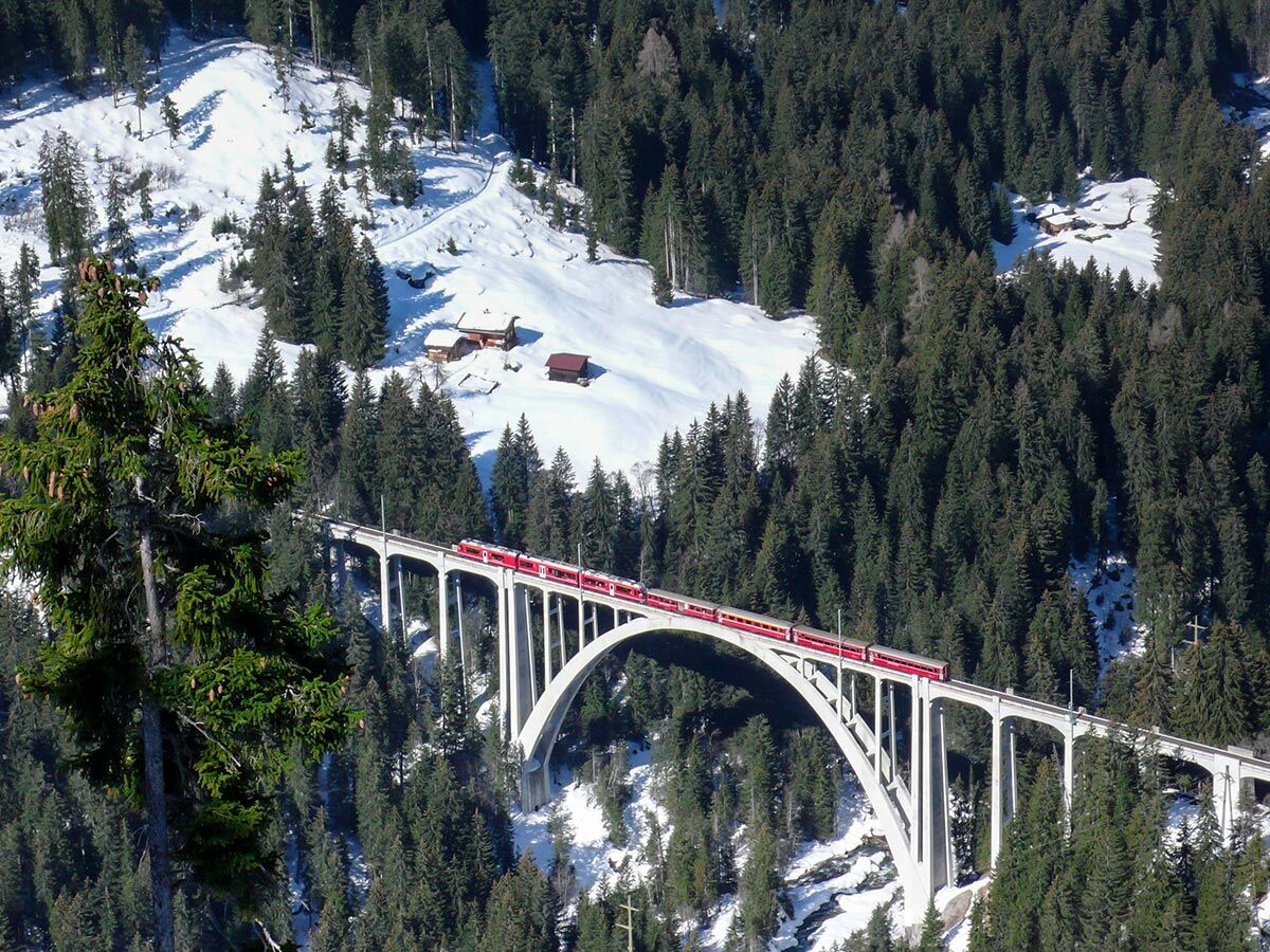 Roter Zug der Rhätischen Bahn fährt über das Landwasserviadukt in verschneiter Winterlandschaft der Schweizer Alpen mit Tannenbäumen.