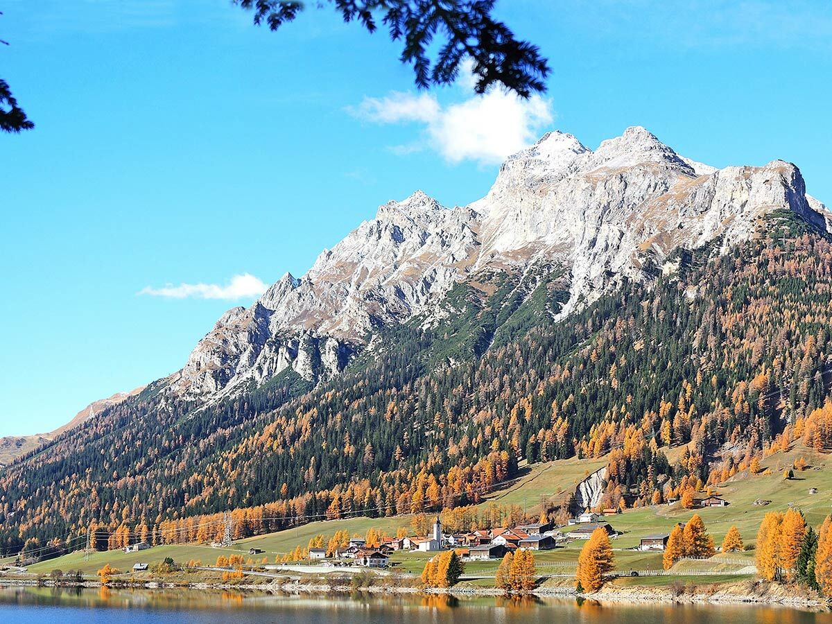 Malerische Herbstlandschaft in den Alpen mit schneebedeckten Bergen, leuchtenden Lärchen, blauem See und kleinem Dorf.