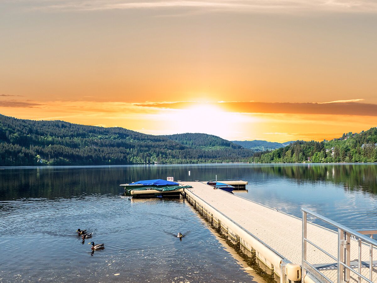 Blick auf einen ruhigen See mit einem langen Steg, schwimmenden Enten und bewaldeten Bergen im warmen Licht des Sonnenuntergangs.