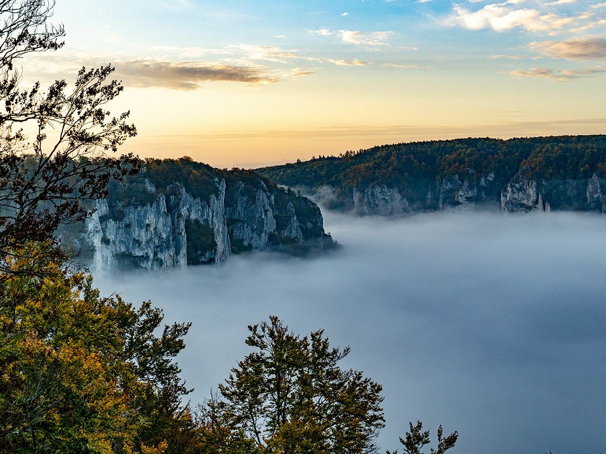 Malerische Berglandschaft mit tiefem Nebel im Tal, umgeben von herbstlichen Bäumen und Felsen bei Sonnenaufgang.