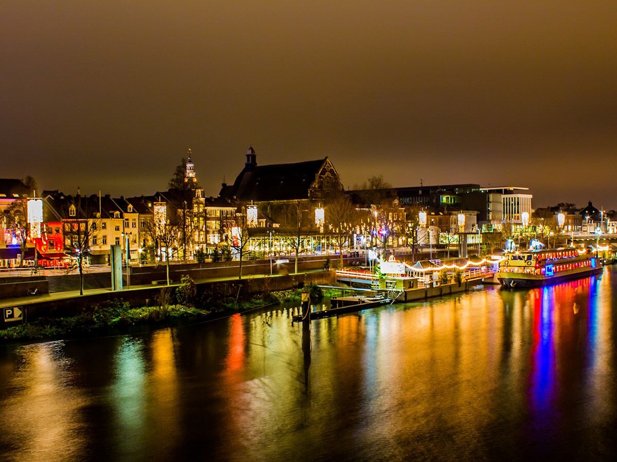 Maastricht bei Nacht: Lebendige Stadtansicht mit beleuchteten historischen Gebäuden, Fluss und bunten Bootsreflexionen auf dem Wasser.