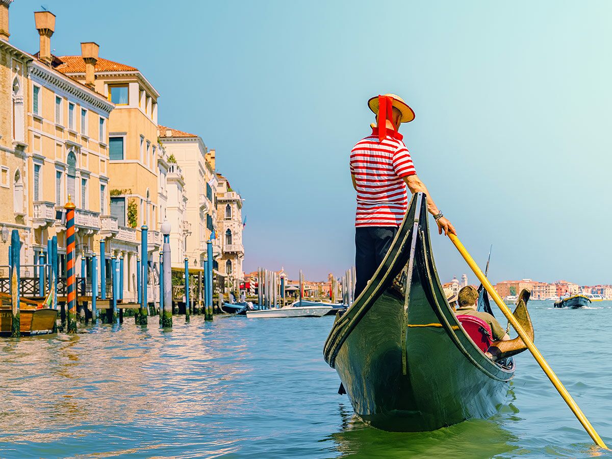 Gondoliere in gestreiftem Shirt rudert Gondel auf Kanal in Venedig mit historischen Gebäuden im Hintergrund bei sonnigem Wetter.