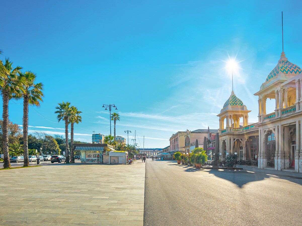 Sonnige Promenade an der italienischen Küste mit Palmen, Jugendstil-Gebäuden, Cafés und Geschäften unter blauem Himmel.