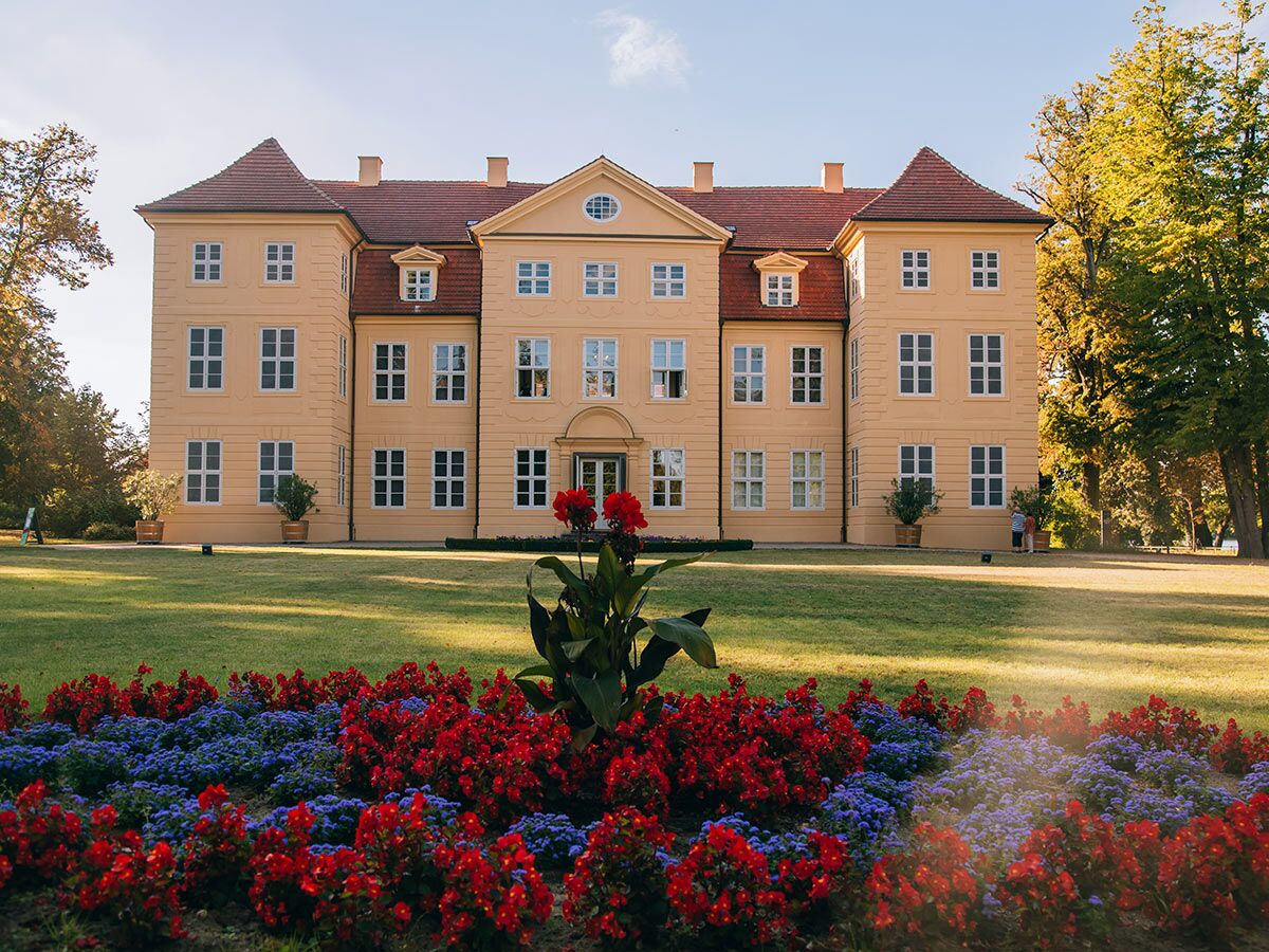 Gelbes, historisches Schloss mit rotem Dach, umgeben von einem grünen Garten mit roten und blauen Blumenbeeten bei Sonnenschein.