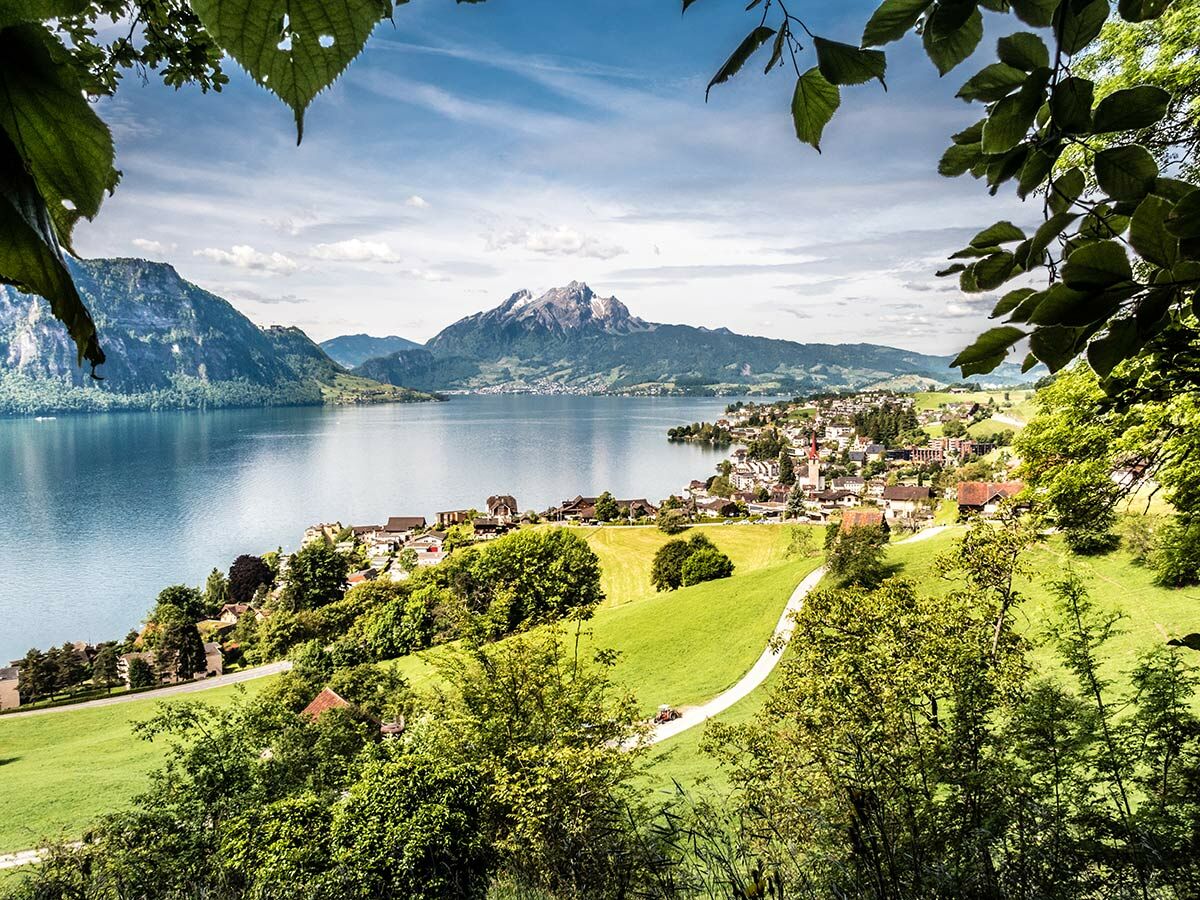 Idyllisches Dorf am See in den Schweizer Alpen, umgeben von grünen Wiesen und Bergen, mit Pilatus im Hintergrund. Laubblätter vorn.