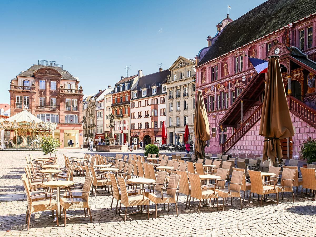 Sonniger europäischer Marktplatz mit leeren Café-Tischen, historischen Gebäuden, Karussell und französischer Flagge.
