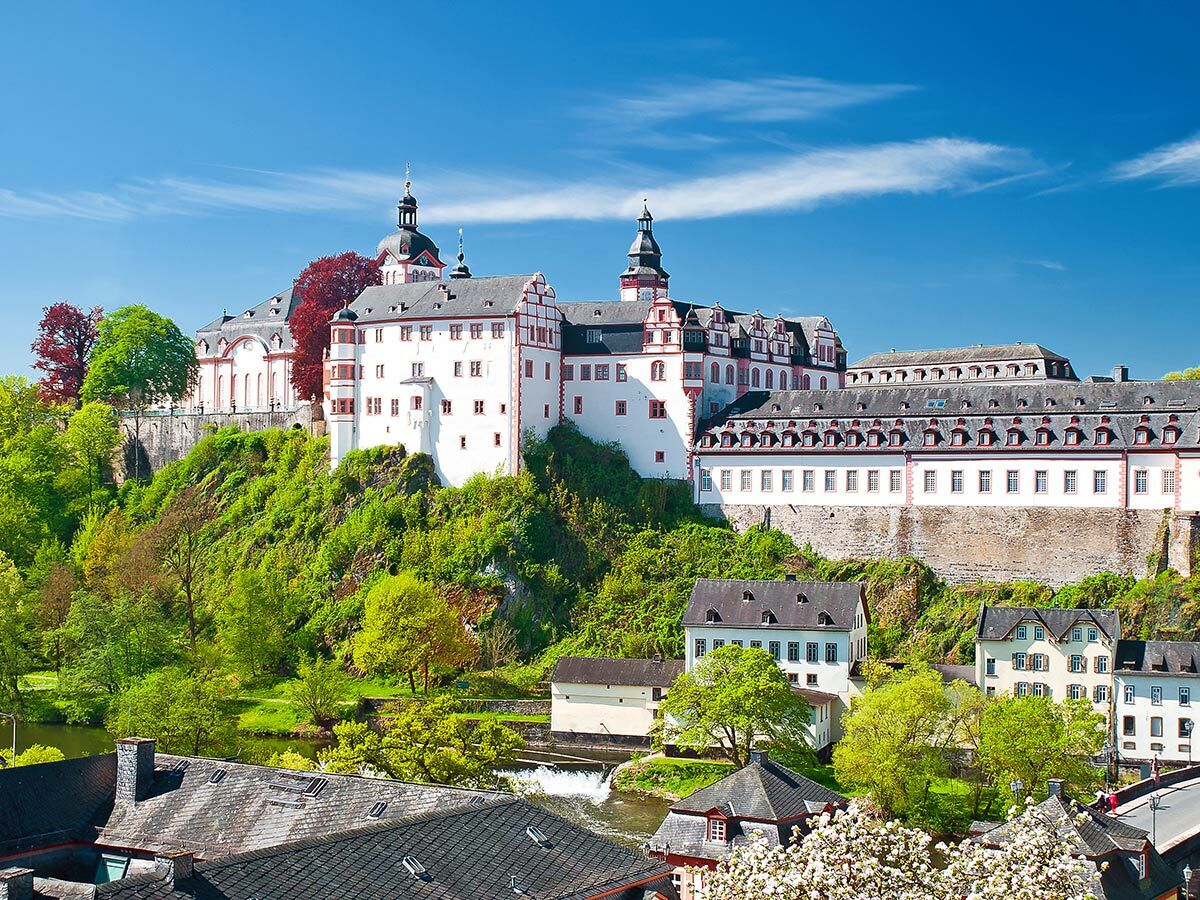 Historische Schlossanlage Weilburg an der Lahn auf grünem Hügel, umgeben von Bäumen und Flusslandschaft unter blauem Himmel.