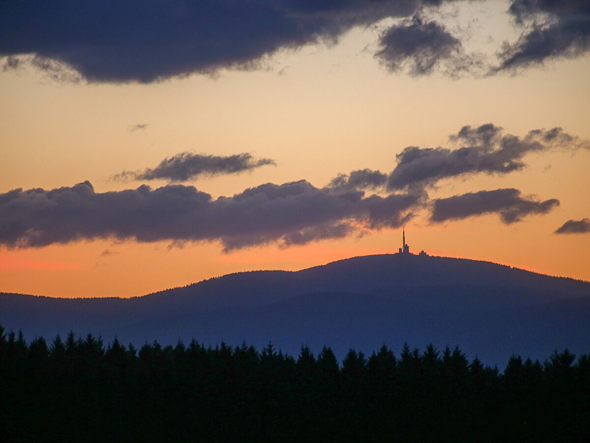 Atmosphärischer Sonnenuntergang über Bergen mit Funkturm auf dem Gipfel, silhouettierte Wälder im Vordergrund und Wolken.