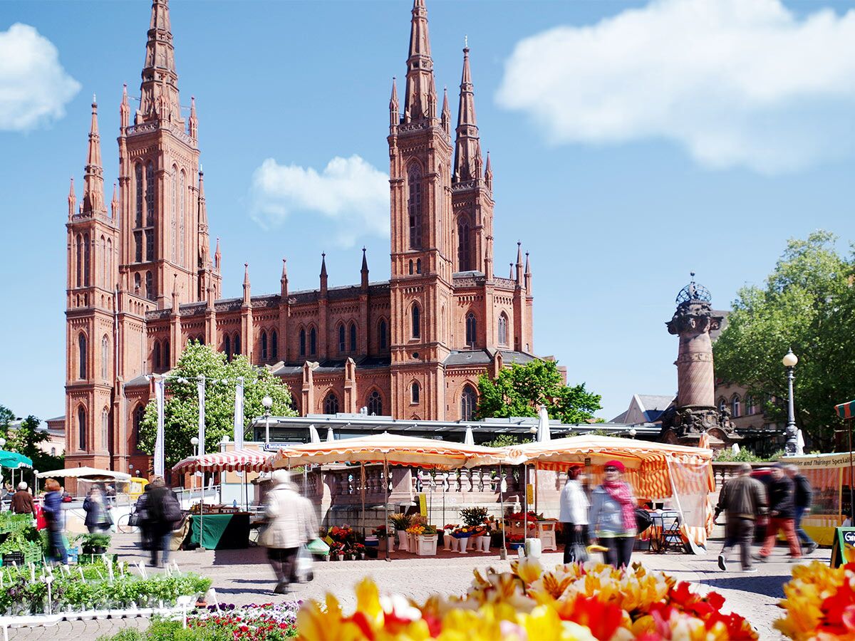 Belebter Wochenmarkt vor der neugotischen Marktkirche in Wiesbaden, mit Blumen und Verkaufsständen unter blauem Himmel.