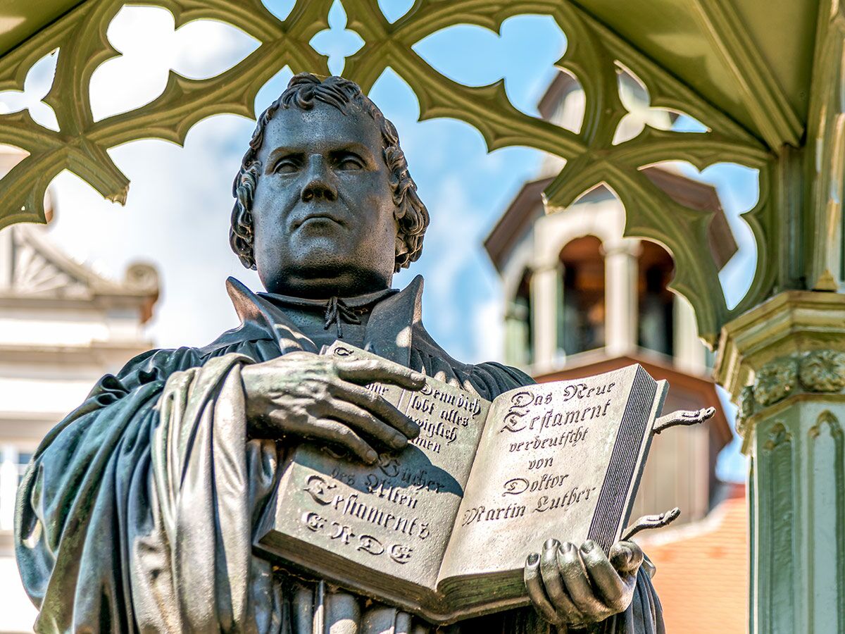 Bronzestatue von Martin Luther mit offenem Buch (Neues Testament) unter einem verzierten Vordach an einem sonnigen Tag.