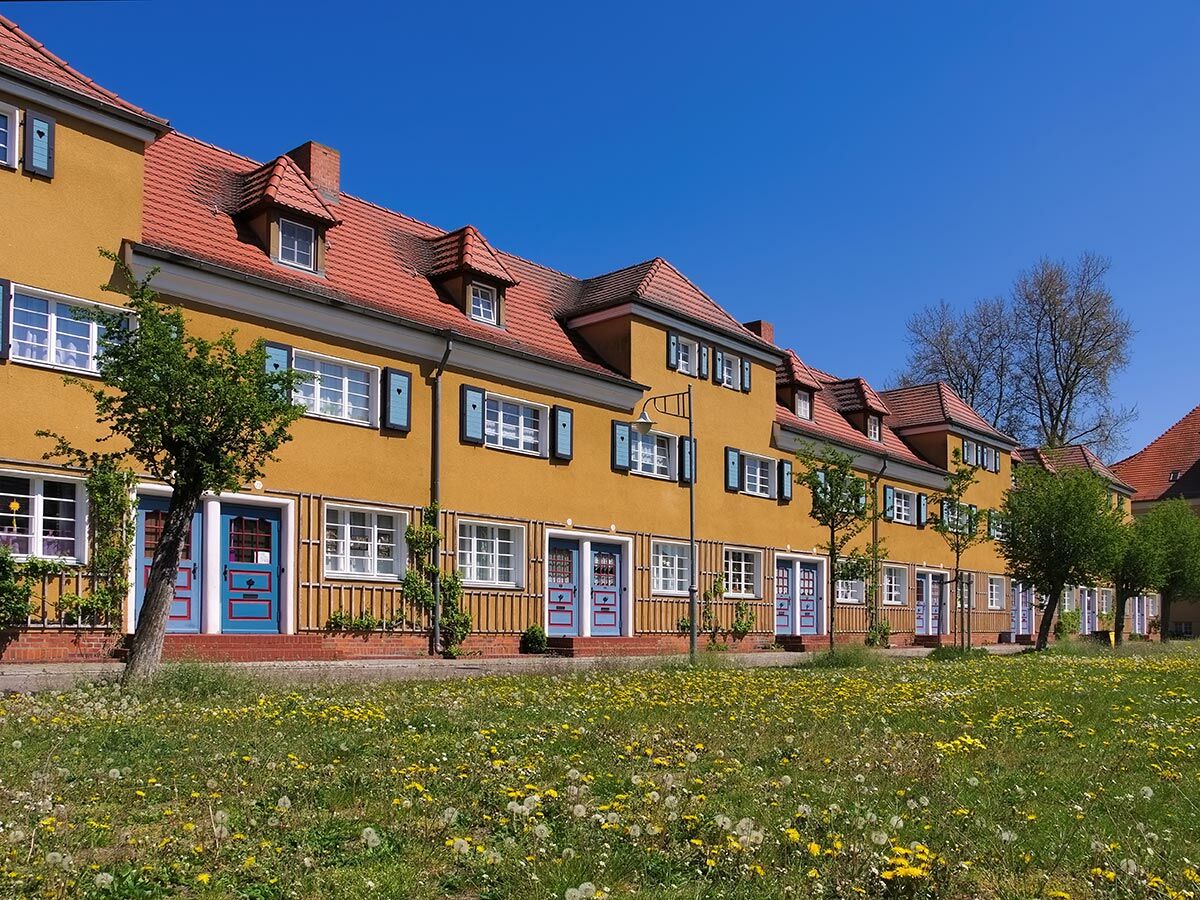 Eine Reihe leuchtend gelber Häuser mit roten Ziegeldächern und blauen Türen, vor einer blühenden Löwenzahnwiese unter blauem Himmel.