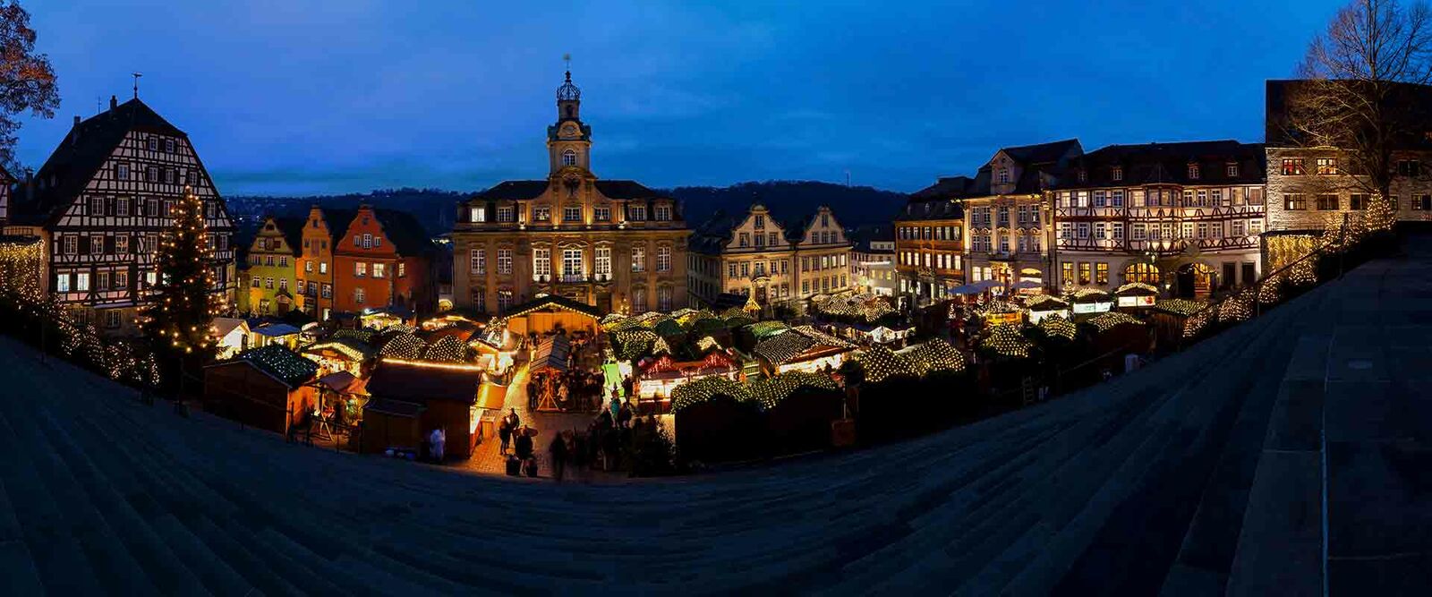 Festlicher Weihnachtsmarkt bei Abenddämmerung mit beleuchteten Ständen und historischen Fachwerkhäusern im Hintergrund.