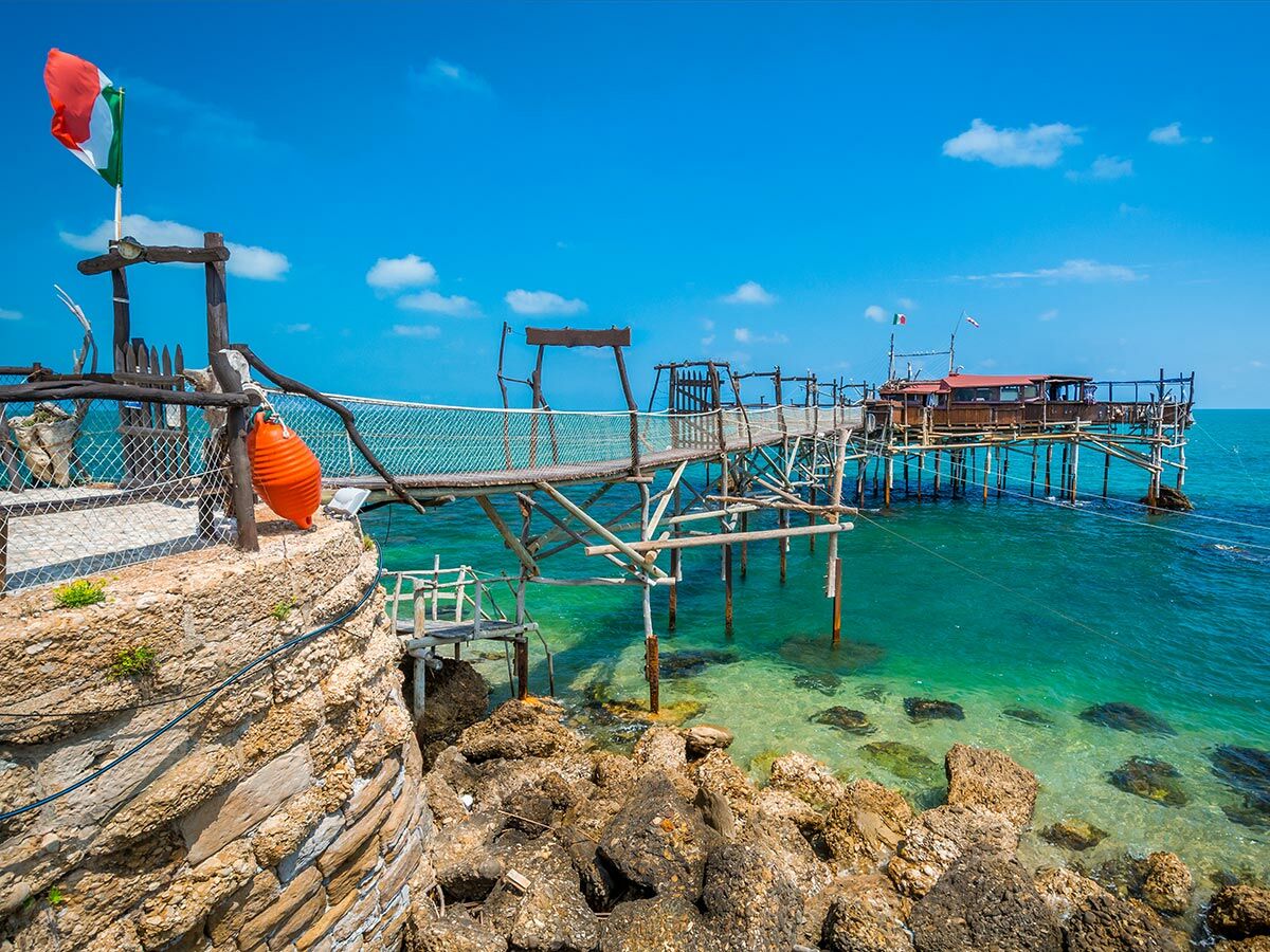 Historischer Trabucco an Italiens Adriaküste. Holzsteg und Fischfangmaschine über türkisfarbenem Meer, Felsen, blauer Himmel, italienische Flagge.