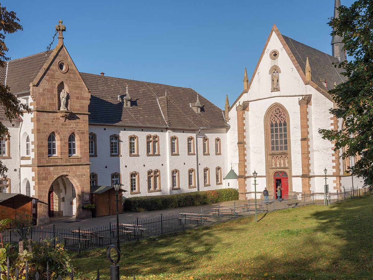 Historisches Kloster Steinfeld mit weißen und steinernen Gebäuden, Kirche, grüner Wiese und Menschen unter blauem Himmel.