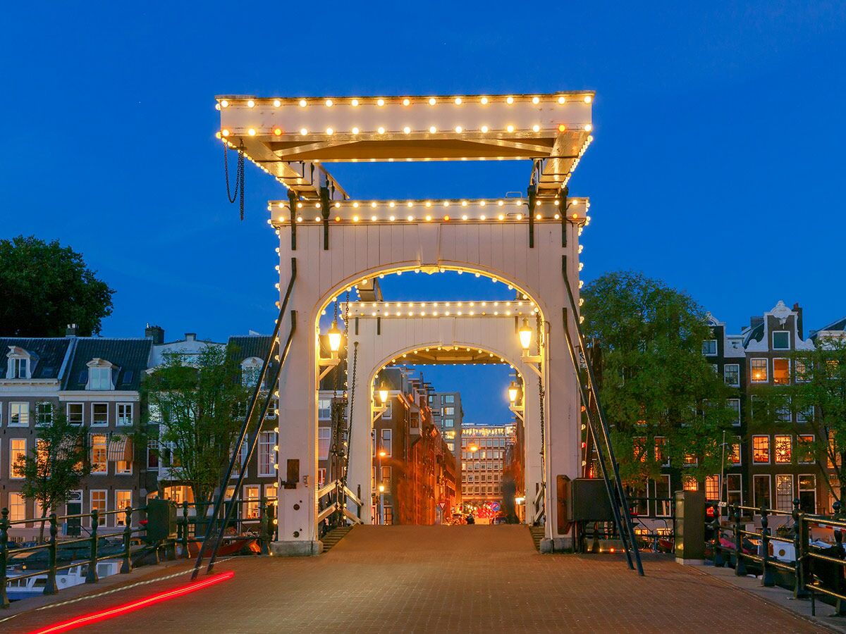 Historische Magere Brug in Amsterdam bei Nacht, weiß beleuchtet über dem Kanal mit traditionellen Häusern. Rote Lichtspur.