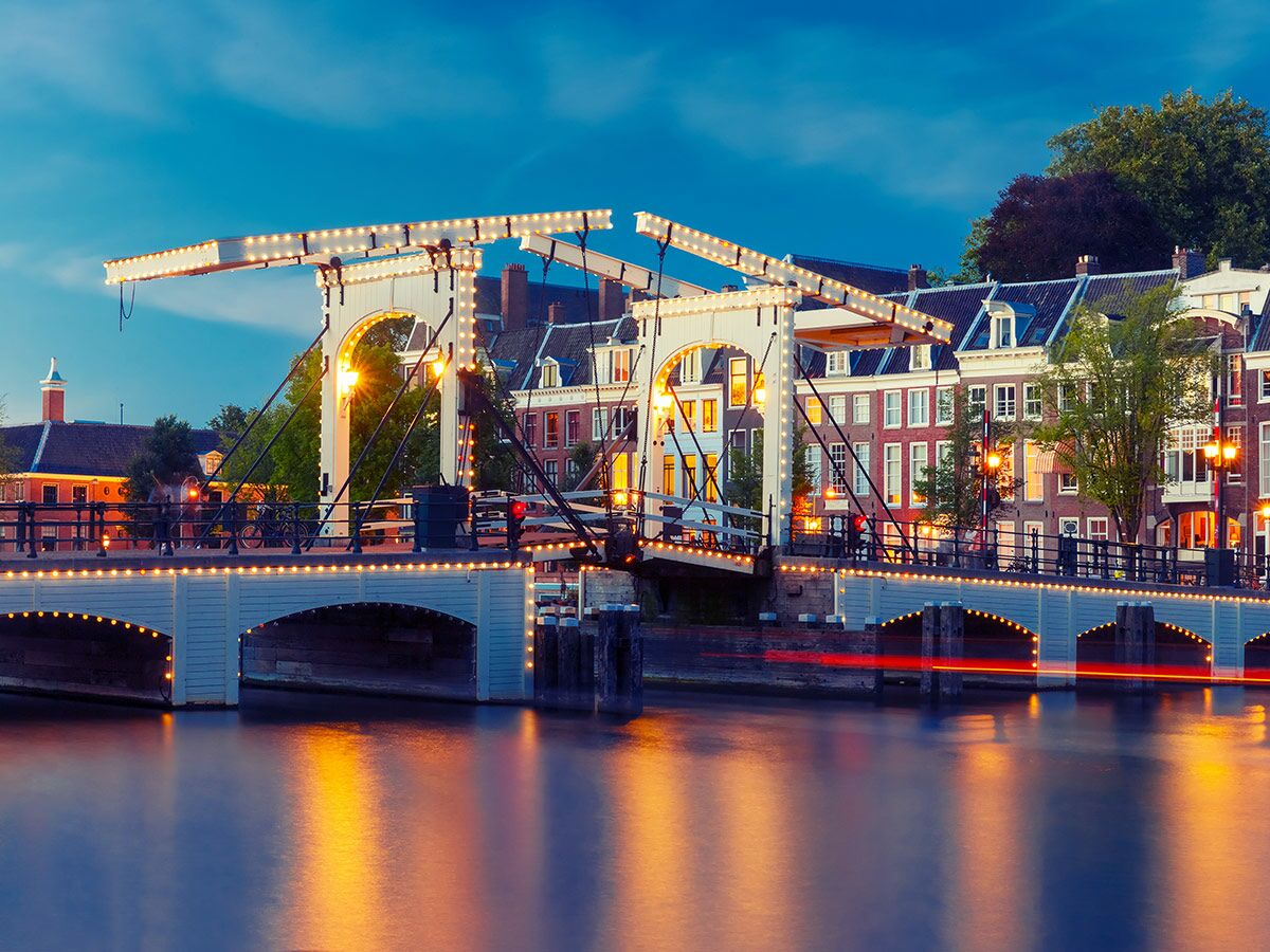 Die beleuchtete Magere Brug (Skinny Bridge) in Amsterdam am Abend mit Reflexionen im Grachtenwasser.