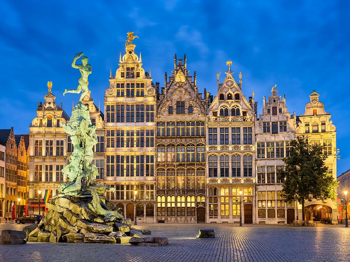 Brabo-Brunnen auf dem Grote Markt in Antwerpen bei Abenddämmerung, umgeben von beleuchteten historischen Zunfthäusern.