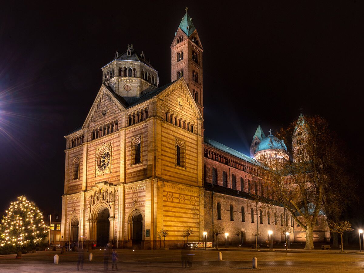 Der Speyerer Dom bei Nacht mit Weihnachtsbaum und warmen Lichtern auf dem Vorplatz, Personen sind verschwommen.
