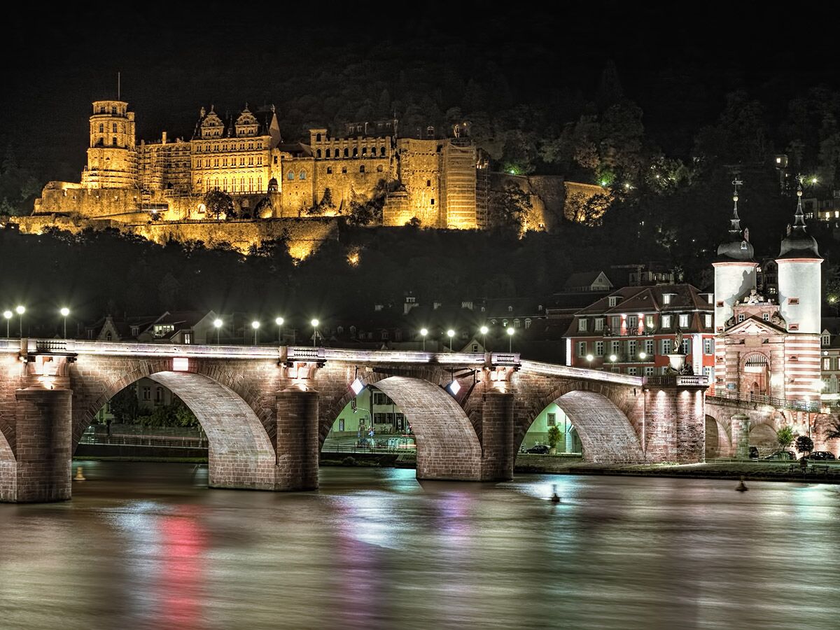 Heidelberg Schloss und Alte Brücke nachts beleuchtet über dem Neckar, mit Brückentor und historischen Gebäuden.