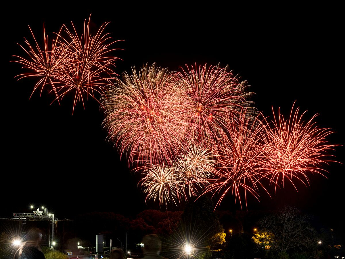 Lebhaftes rotes und goldenes Feuerwerk erhellt den dunklen Nachthimmel über einer beleuchteten Stadtansicht mit Zuschauern.