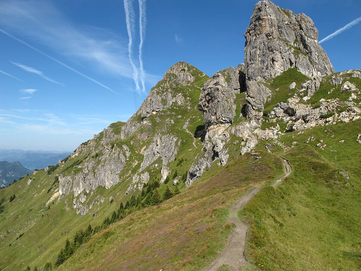 Panoramablick auf einen gewundenen Bergpfad in den grünen Alpen, flankiert von hohen Felsgipfeln unter blauem Himmel mit Kondensstreifen.