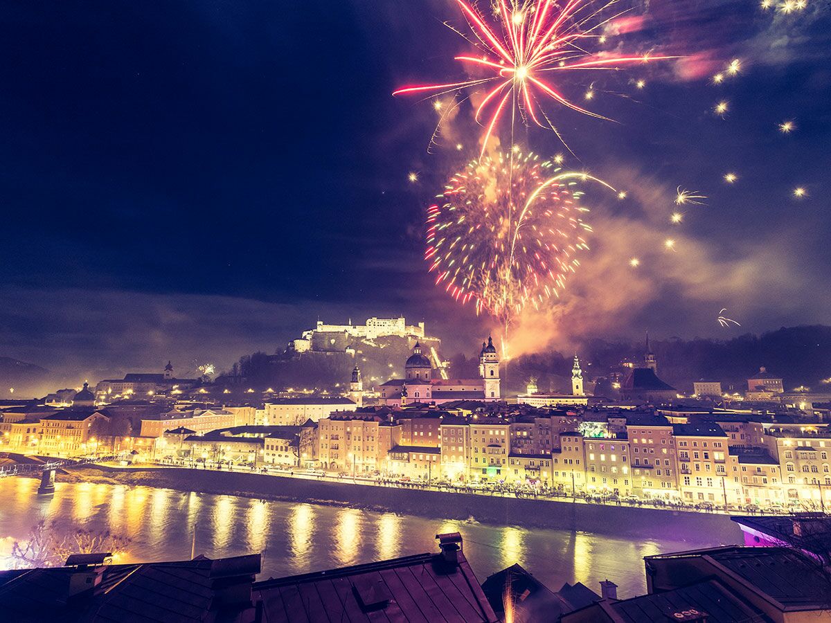 Festliches Feuerwerk über der beleuchteten Altstadt von Salzburg mit der Festung Hohensalzburg und dem Fluss Salzach bei Nacht.