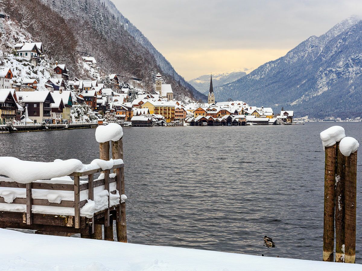 Idyllisches Hallstatt Winterdorf in Österreich mit schneebedeckten Häusern, See, Bergen, Kirche und Holzsteg im Vordergrund.