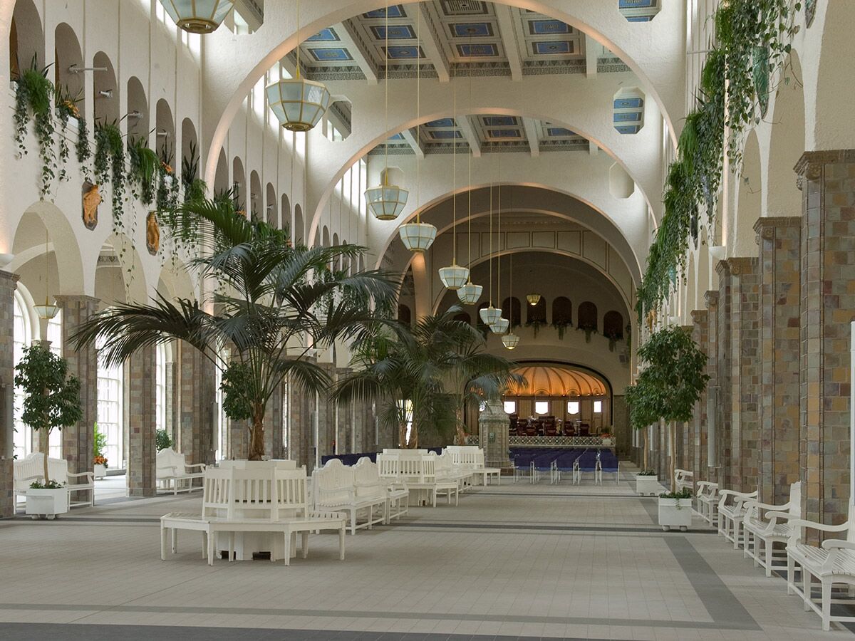 Geräumige, helle Innenhalle mit hohen Bögen, Palmen, weißen Sitzbänken und eleganten Kronleuchtern. Hängepflanzen schmücken Wände.