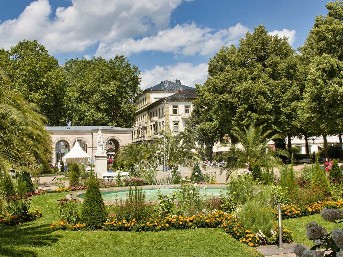Eine lebhafte Parkszenen mit spritzendem Springbrunnen, farbenfrohen Blumen, saftigem Grün und historischem Gebäude unter blauem Himmel.