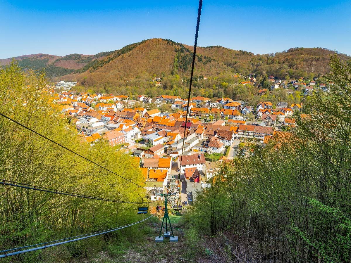 Blick vom Sessellift auf eine malerische Stadt mit orangefarbenen Dächern im Harz, umgeben von grünen Hügeln und Wäldern unter blauem Himmel.
