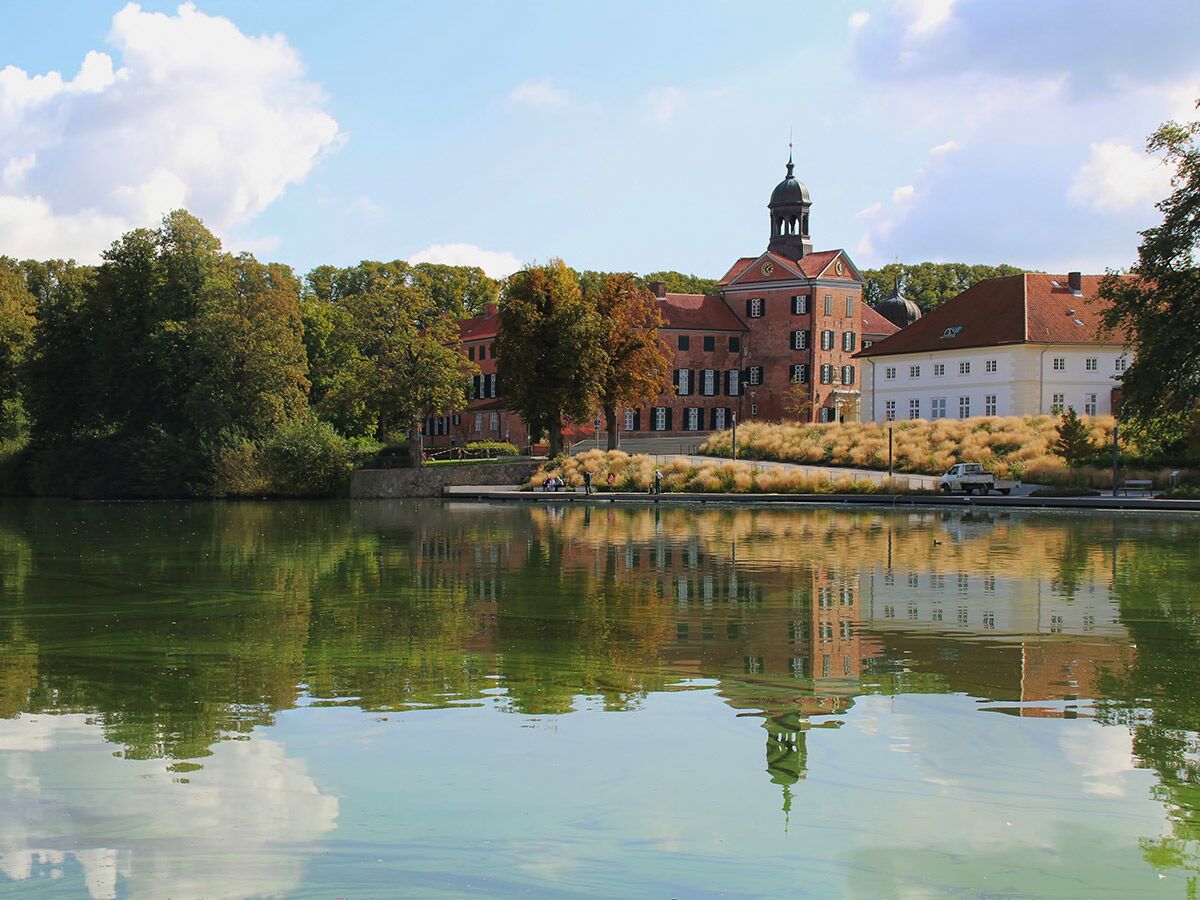 Historisches Schloss am Seeufer, umgeben von herbstlichen Bäumen, mit klarer Spiegelung im Wasser und blauem Himmel.