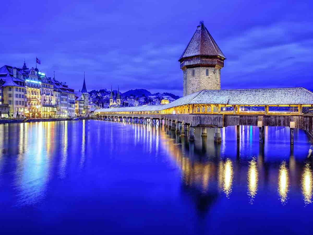 Historische Kapellbrücke und Wasserturm in Luzern, Schweiz, im Winter bei Nacht. Schnee bedeckt Dächer, Lichter spiegeln sich im Fluss.