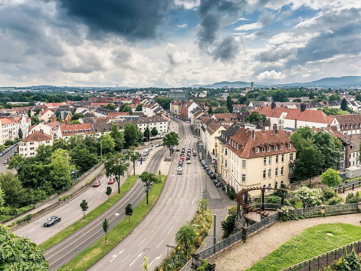 Panoramablick auf ein europäisches Stadtbild mit kurviger Straße, vielen Häusern, roten Dächern, grünen Bäumen und Bergen unter bewölktem Himmel.