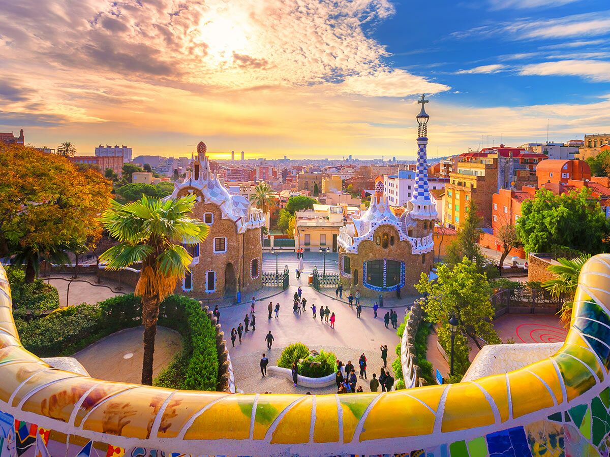 Panoramablick auf Park Güell Barcelona mit Gaudís Pförtnerhäusern, Mosaiken und Stadt bei Sonnenuntergang.