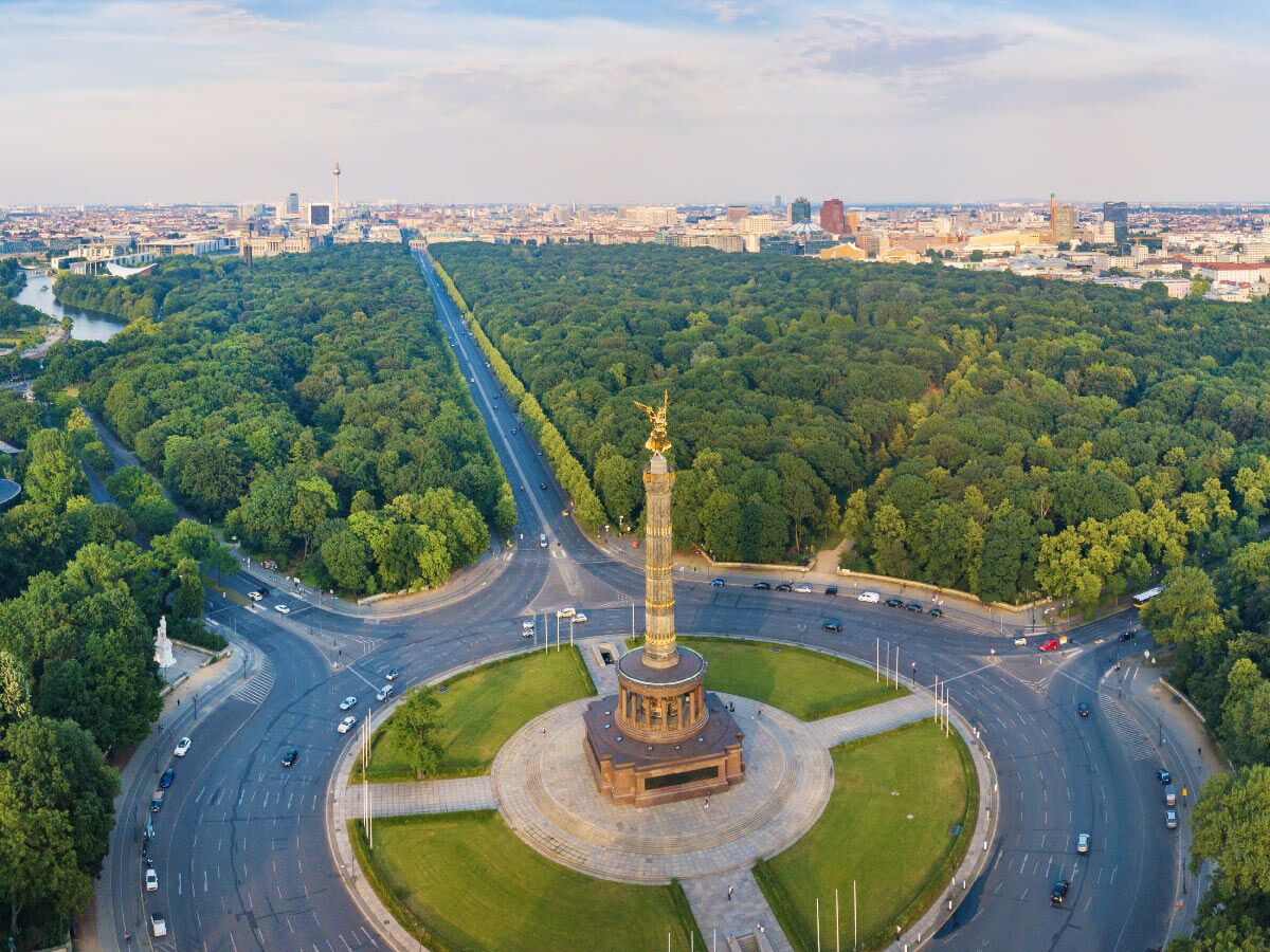 Luftbild der Siegessäule in Berlin mit dem Tiergarten, Straßenverkehr und der Stadtkulisse im Hintergrund bei sonnigem Wetter.