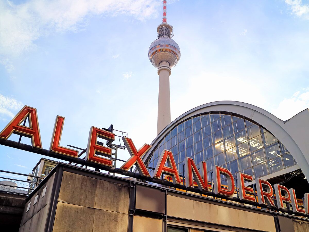 Rotes "ALEXANDERPLATZ"-Schild vor dem Berliner Fernsehturm und einem Gebäude mit Glasfassade bei Tag.