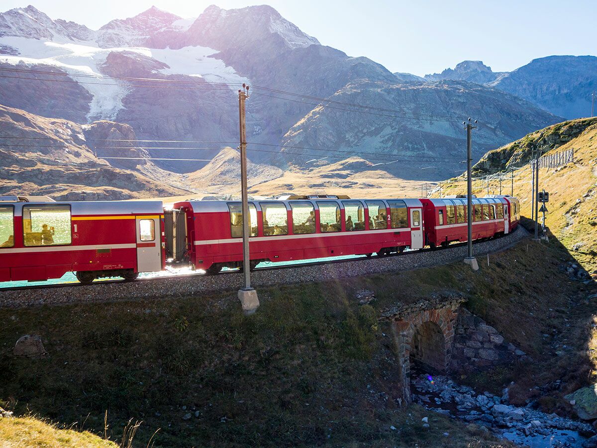 Roter Panoramazug fährt durch alpine Berglandschaft mit Gletscher und sonnigem Himmel.