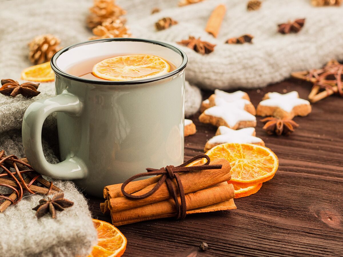Gemütliche Wintertee-Tasse mit Orangenscheibe, Zimt, Sternanis und Weihnachtsplätzchen auf einer Strickdecke und Holztisch.