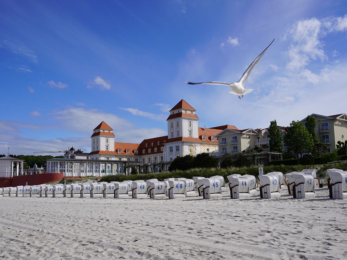 Elegantes Hotel an der Ostsee mit Strandkörben und fliegender Möwe am weißen Sandstrand unter blauem Himmel.