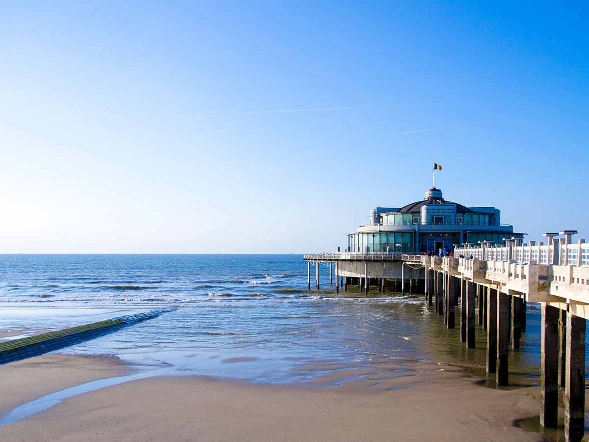 Blick auf den historischen Blankenberge Pier mit dem runden Gebäude am Ende, das ins ruhige Meer hineinragt, und Sandstrand im Vordergrund unter blauem Himmel.