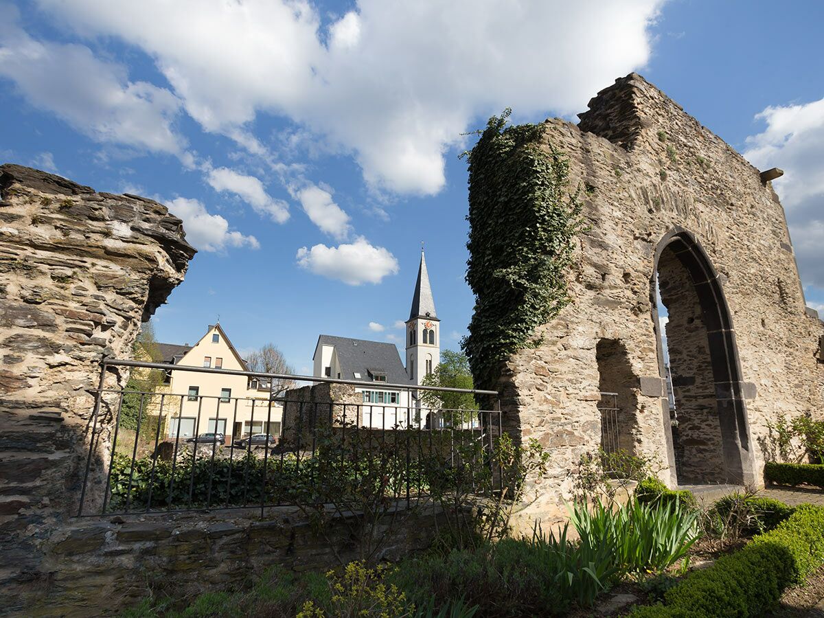 Alte Steinruine mit Torbogen und Efeu, im Hintergrund eine Dorfkirche mit spitzem Turm und Häuser unter blauem Himmel mit Wolken.