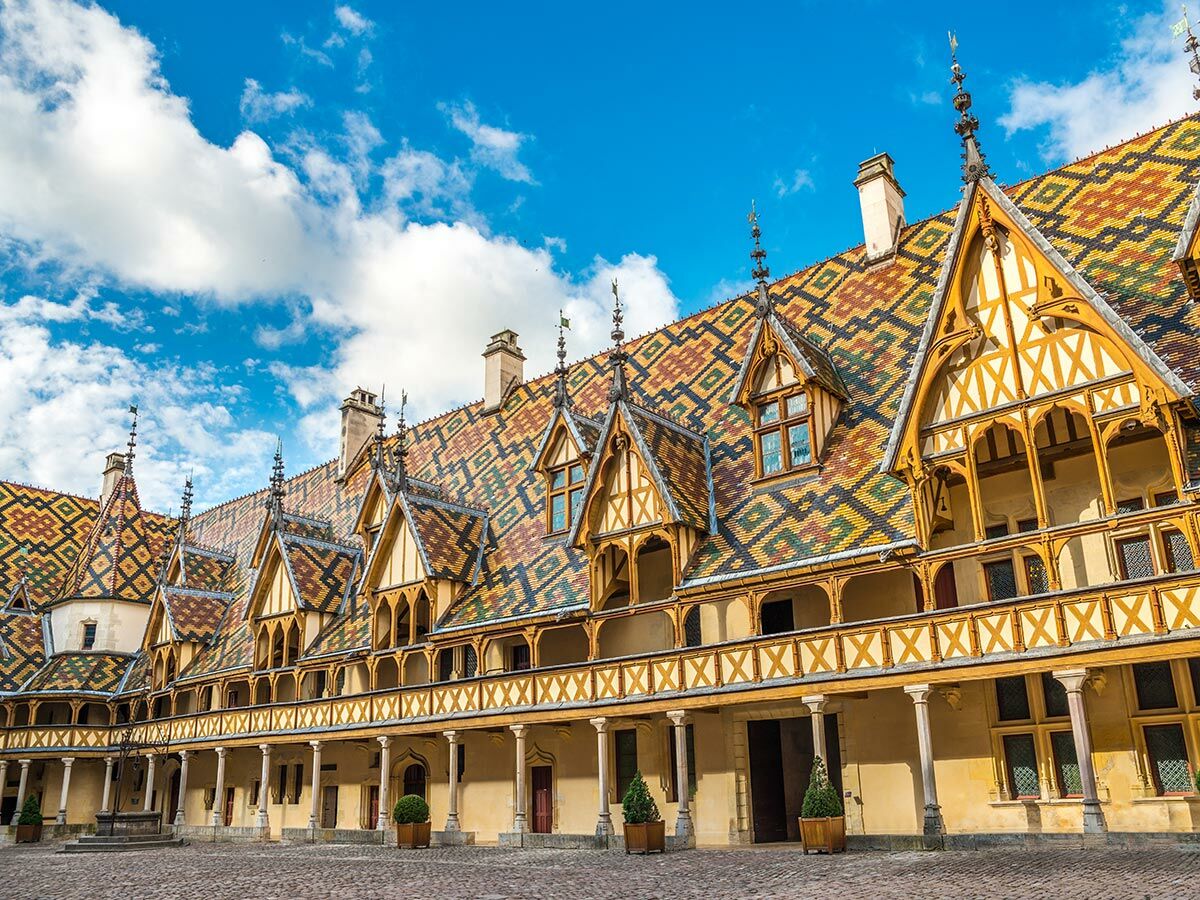 Hospices de Beaune in Burgund mit ikonischem, farbigem Ziegeldach und Innenhof unter blauem Himmel.