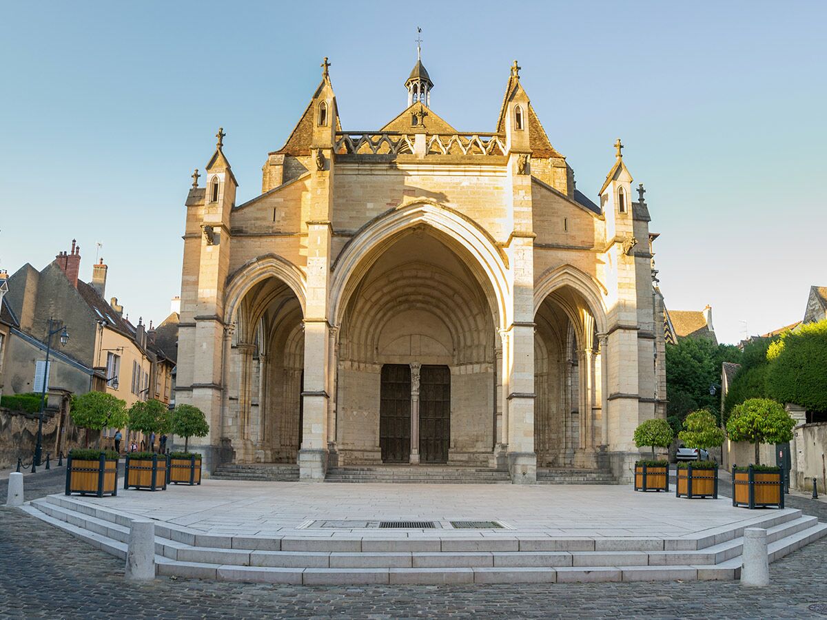 Die Fassade einer historischen Steinkathedrale mit drei Bögen und kleinen Bäumen auf einem gepflasterten Platz bei Sonnenuntergang.