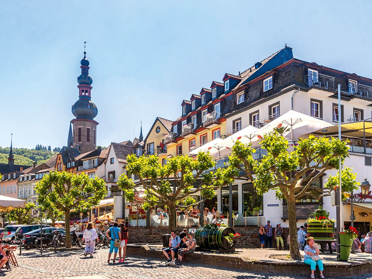 Lebhafter Marktplatz in Cochem an der Mosel mit Kirche, historischen Gebäuden, Menschen und Cafés unter blauem Himmel.