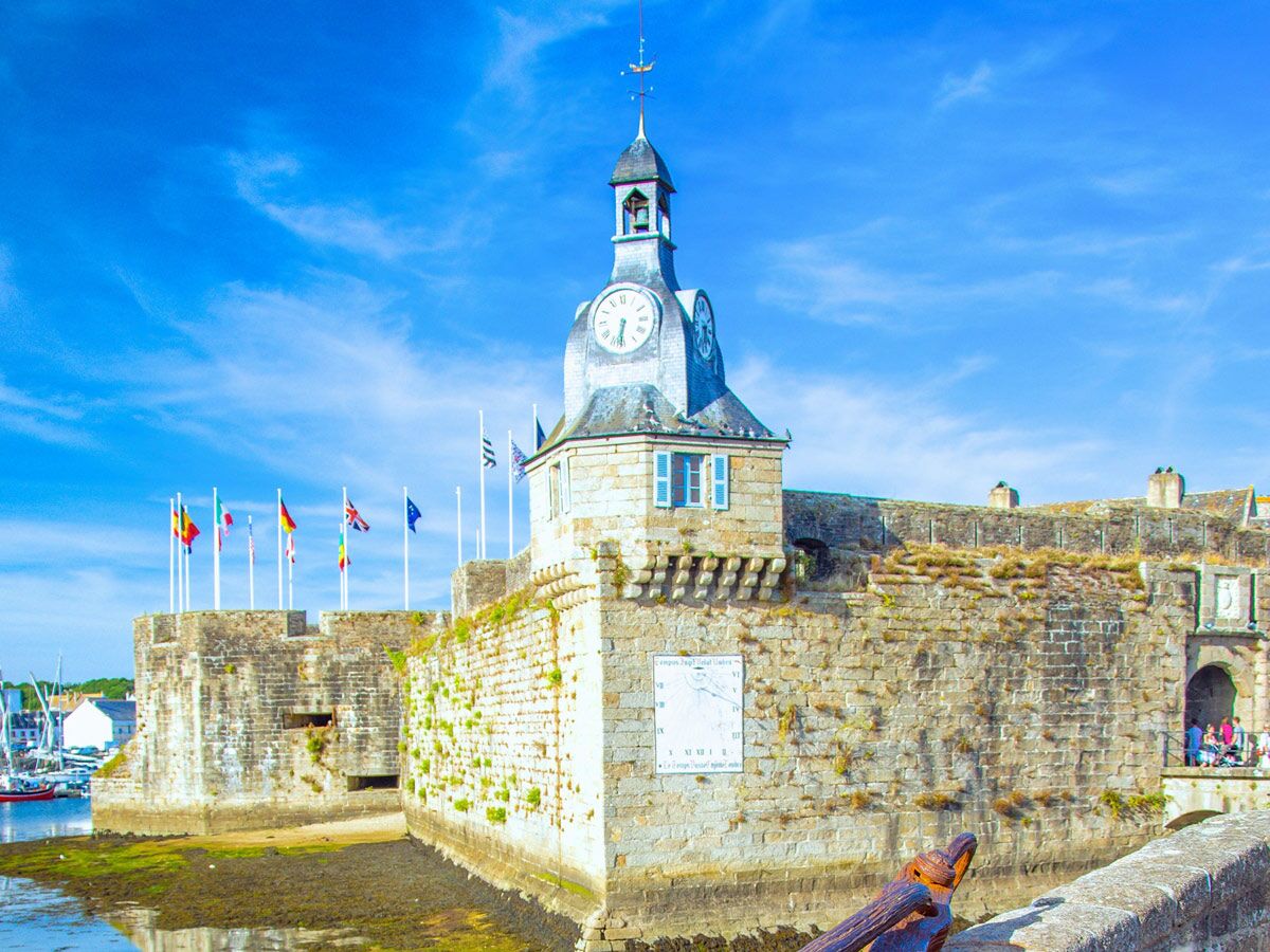 Panoramabild der historischen Festung Concarneau mit Uhrturm, Sundial, Flaggen und Hafen unter blauem Himmel.