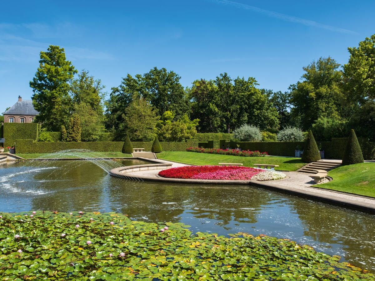 Wunderschöner formaler Garten mit großem Teich, Seerosen, Springbrunnen, bunten Blumenbeeten und Formschnittgehölzen unter blauem Himmel.