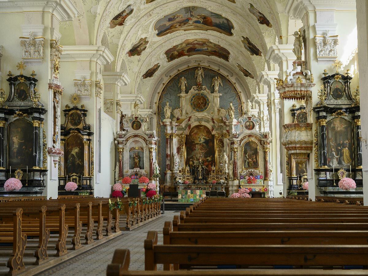 Prächtiger barocker Kircheninnenraum mit bemalter Decke, goldenen Verzierungen, kunstvollen Altären und Holzbänken.