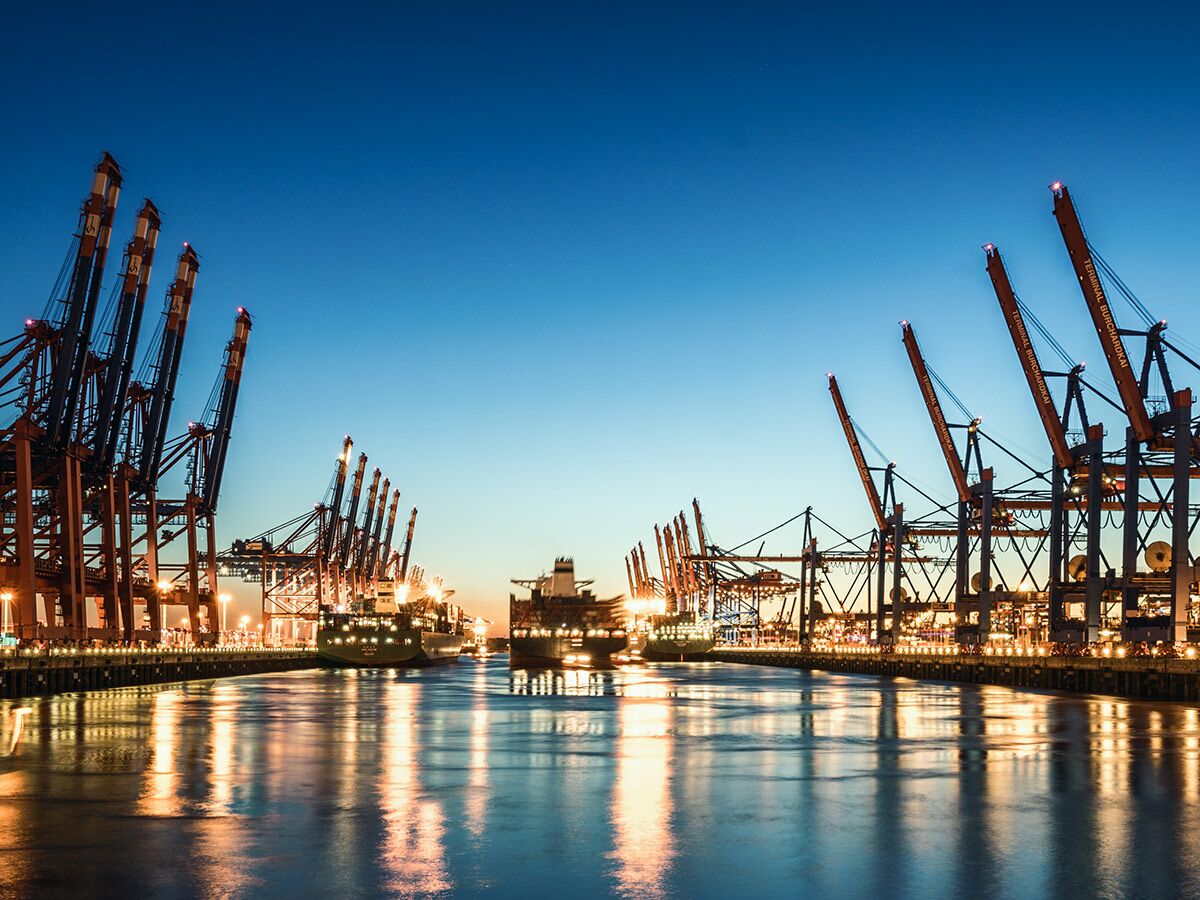 Beleuchtete Containerkräne und Frachtschiffe im Hamburger Hafen bei Dämmerung, mit Lichtreflexionen auf dem Wasser des Burchardkais.