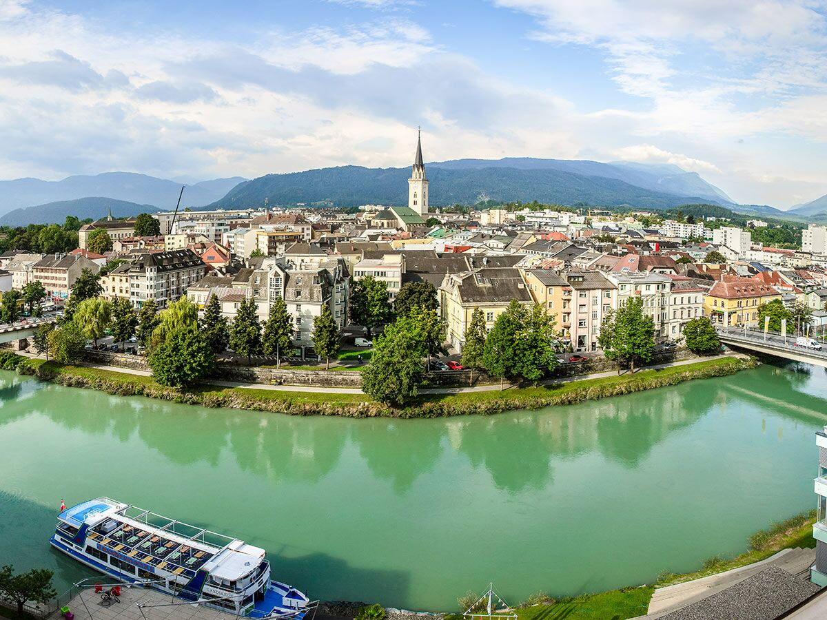 Panorama von Villach mit der Drau, grünen Bäumen, historischen Gebäuden und einem Flusskreuzfahrtschiff vor Bergkulisse.