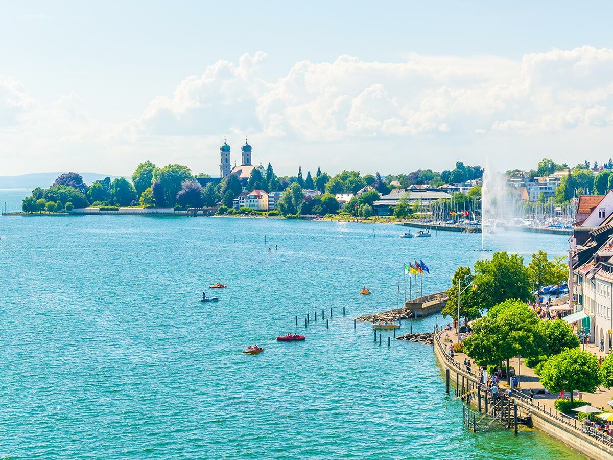 Panoramablick auf Konstanz am Bodensee mit Münster, Hafen, Promenade und Menschen bei Wassersport unter blauem Himmel.